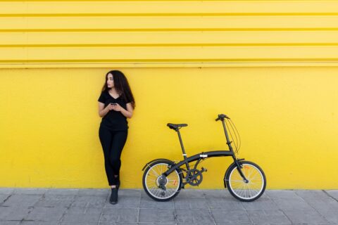 Woman dressed in black standing in front of a yellow wall next to a black folding bike